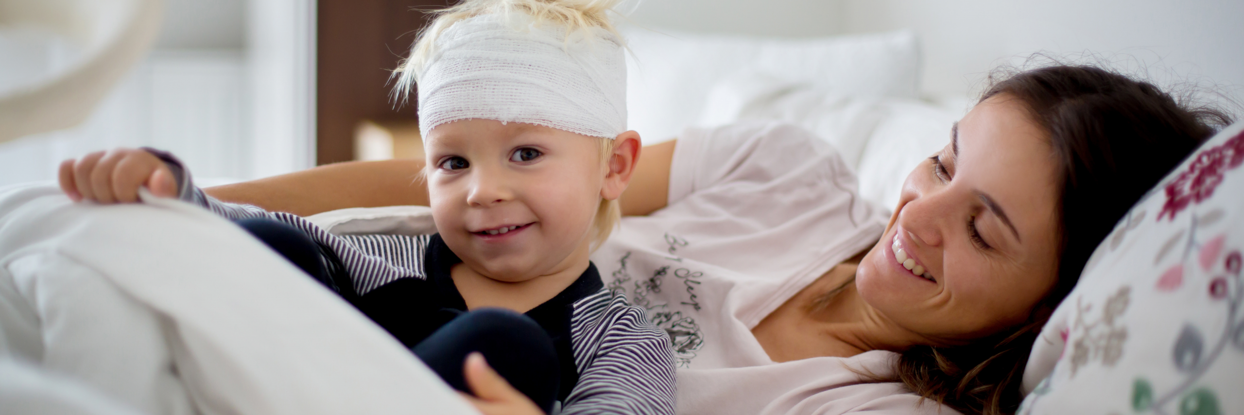 Little boy with a bandage around his head sitting in a bed with his mom laying down. Both are smiling.