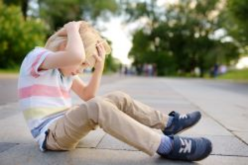 Blonde little boy sitting on sidewalk and holding his head with both of his hands