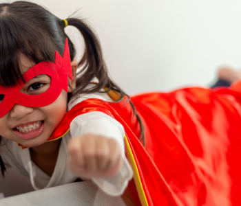 Dark-haired little girl laying on back of couch dressed as a superhero with her arms outstretched